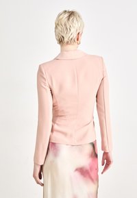 Lys rosa, tettsittende blazer med lange ermer, hakkekrage og rene linjer. Glatt stofftekstur uten synlige mønstre eller pynt.