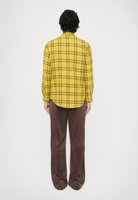 Żółta koszula w kratę z długimi rękawami i mankietami na guziki, zestawiona z luźnymi brązowymi spodniami. Koszula ma wysoki kołnierz i zaokrąglony dół.
