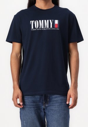 Homme portant un t-shirt bleu marine avec le logo Tommy blanc et rouge, accompagné du texte "Tommy Jeans Worldwide Since 1985", porté avec un jean bleu.