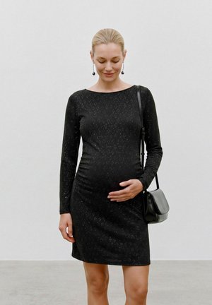 Femme enceinte en robe noire à manches longues, tenant doucement son ventre d'une main, portant des boucles d'oreilles et un sac à épaule.