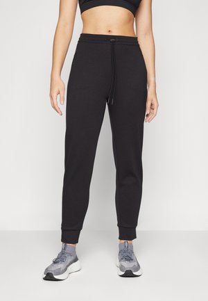 Frau trägt schwarze Sweatpants mit Kordelzug und graue Sportschuhe, steht vor einem einfarbigen hellen Hintergrund.