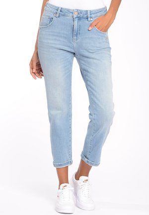 Nainen pukeutuneena vaaleansinisiin käännettyihin farkkuihin ja valkoisiin Nike-tennareihin, seisoo käsi taskussa valkoista taustaa vasten.