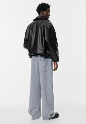 Čierna kožená bunda s voľným strihom a širokým golierom, skombinovaná so šedými teplákmi so bočnou vreckom. Pohľad zozadu.
