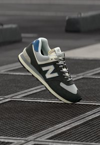 New Balance 574 sneaker, szürke velúr és hálós felsőrésszel, kék kiegészítőkkel és fekete gumitalppal, egy texturált felületen.