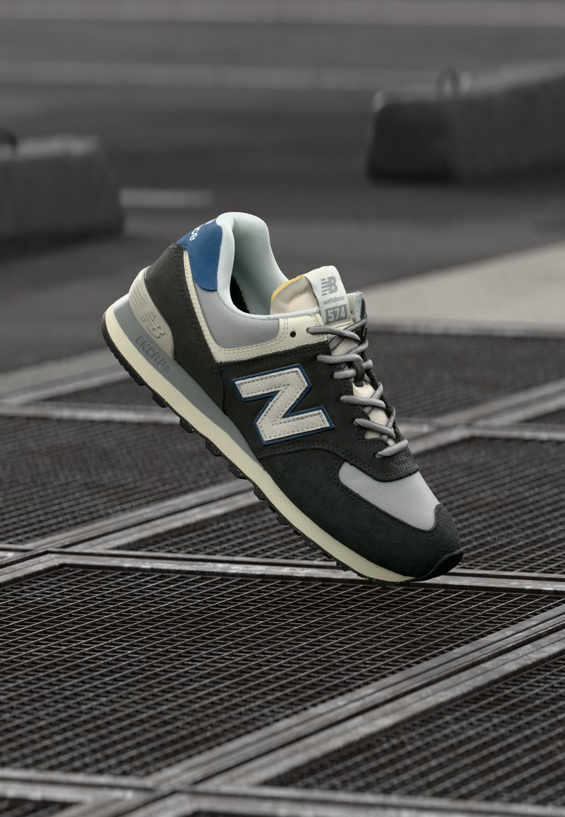 New Balance 574 sneaker, szürke velúr és hálós felsőrésszel, kék kiegészítőkkel és fekete gumitalppal, egy texturált felületen.
