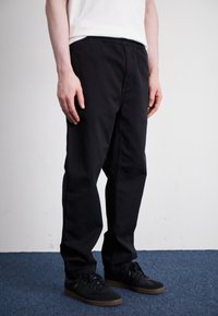 Black cotton trousers featuring an elastic waistband, straight fit, and side pockets, paired with black sneakers on a blue carpet.
