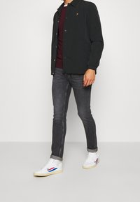 Schwarze leichte Jacke mit goldenem Emblem, burgunderrotes T-Shirt mit Rundhalsausschnitt, graue Slim-Fit-Jeans und weiße Sneaker mit roten und blauen Akzenten.