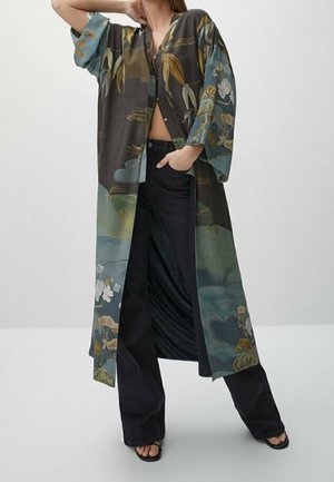 Donna che indossa una lunga giacca stile kimono a fiori sopra pantaloni neri a gamba larga e sandali neri con punta aperta, posando con una mano sui fianchi.