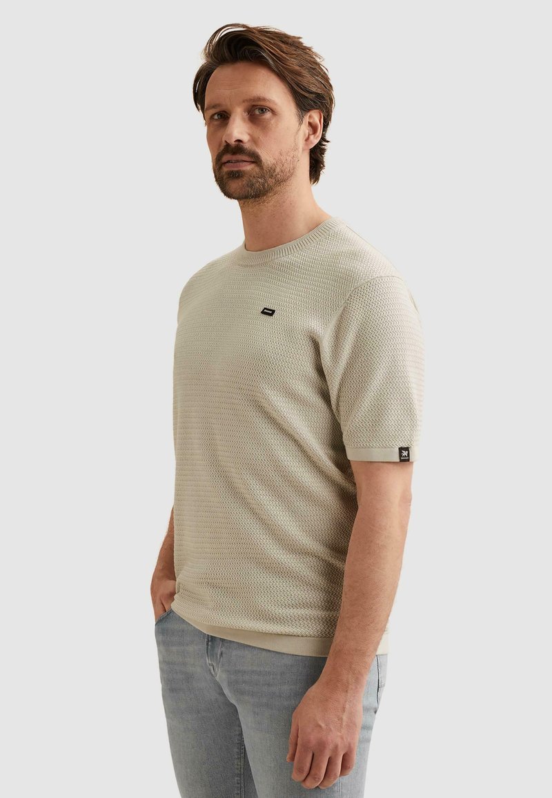 Homme portant un pull beige texturé à manches courtes et un jean bleu clair, debout devant un fond gris clair uni.