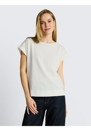 Femme blonde portant un haut blanc texturé à manches courtes et un jean foncé, debout avec un léger sourire sur un fond blanc uni.