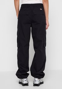 Personne portant un pantalon cargo noir avec taille élastique et chaussures de sport argentées, vue de dos sur un fond uni.