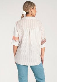 Pasywny widok z tyłu koszuli w kolorze białym i jasnoróżowym, z haftowanymi wzorami kwiatowymi na rękawach oraz luźnym krojem.