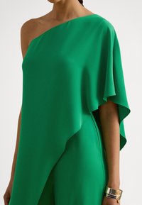 Vestido verde de um ombro só, em tecido leve, com uma caída fluida e bainha assimétrica. Acompanhado por uma pulseira dourada.