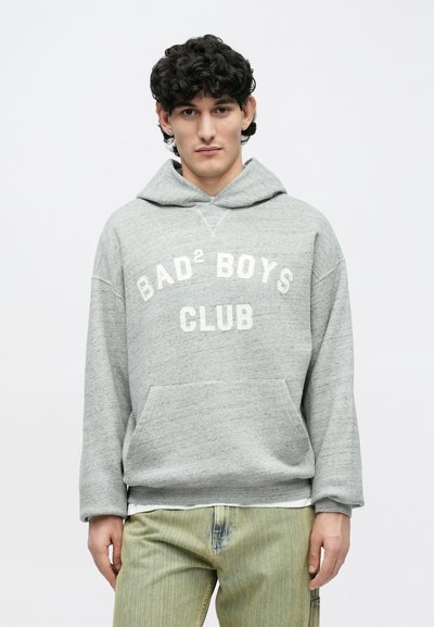 Giovane uomo con capelli ricci scuri indossa una felpa grigia con il testo "BAD² BOYS CLUB" e pantaloni verde chiaro, in piedi davanti a uno sfondo bianco uniforme.