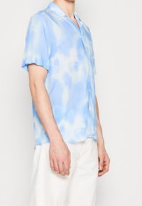 Kortärmad skjorta i ljusblått med mönster i tie-dye, gjord av mjukt tyg. Har en krage och knäppning med knappar.