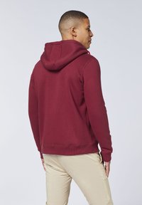 Burgunderfarbene Kapuzensweatshirt aus weichem Stoff, mit einer Vorderlasche, gerippten Bündchen und einer lässigen Passform. Getragen mit hellen Hosen.