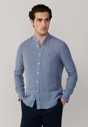 Junger Mann, der ein hellblaues Hemd mit Stehkragen und langen Ärmeln trägt, steht mit den Händen in den Taschen seiner marineblauen Hose und blickt nach rechts.