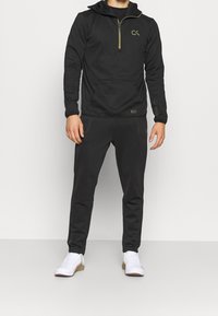 Felpa nera con zip e dettagli verde, tasca frontale e pantaloni aderenti coordinati. Indossata con scarpe sportive bianche. Design semplice e funzionale.