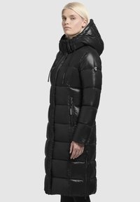 Black puffer coat featuring a long, fitted design, quilted texture, large hood, and zippers on side pockets. Made from a shiny material.
