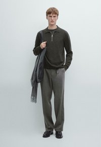 Maglione a maglia grigio scuro con collo a zip, pantaloni grigi su misura e scarpe nere. Il modello tiene in mano una sciarpa grigia e una borsa. Sfondo neutro.