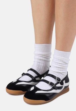 Beine, die weiße gerippte Socken tragen, kombiniert mit schwarz-weißen Leder-Mary Jane Schuhen mit doppelten Schnallenriemen und Gummisohlen.