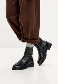 Persona que lleva pantalones marrones arremangados, calcetines de canalé grises y botas de cuero negras hasta el tobillo con cremalleras laterales y tacones bajos.