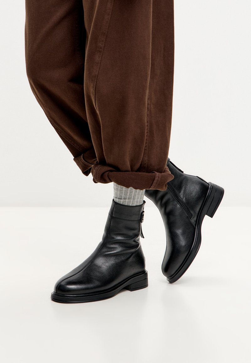 Persona que lleva pantalones marrones arremangados, calcetines de canalé grises y botas de cuero negras hasta el tobillo con cremalleras laterales y tacones bajos.