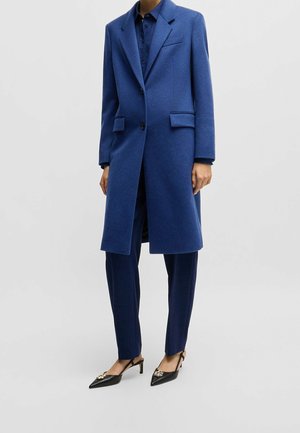 Vrouw draagt een lange blauwe wollen jas over bijpassende blauwe broek en blouse, gecombineerd met zwarte slingback hakken met puntige neus en gouden versieringen.