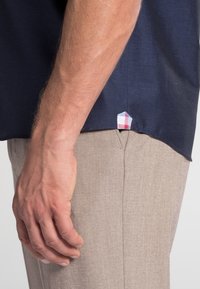 Chemise à manches courtes bleu marine avec une texture subtile, associée à un pantalon beige clair. Détail notable : une petite étiquette à carreaux au bas de la chemise.