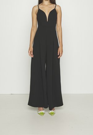 Femme portant une combinaison noire sans manches à jambes larges et col en V profond, associée à des chaussures vertes pointues ornées de nœuds en cristal.