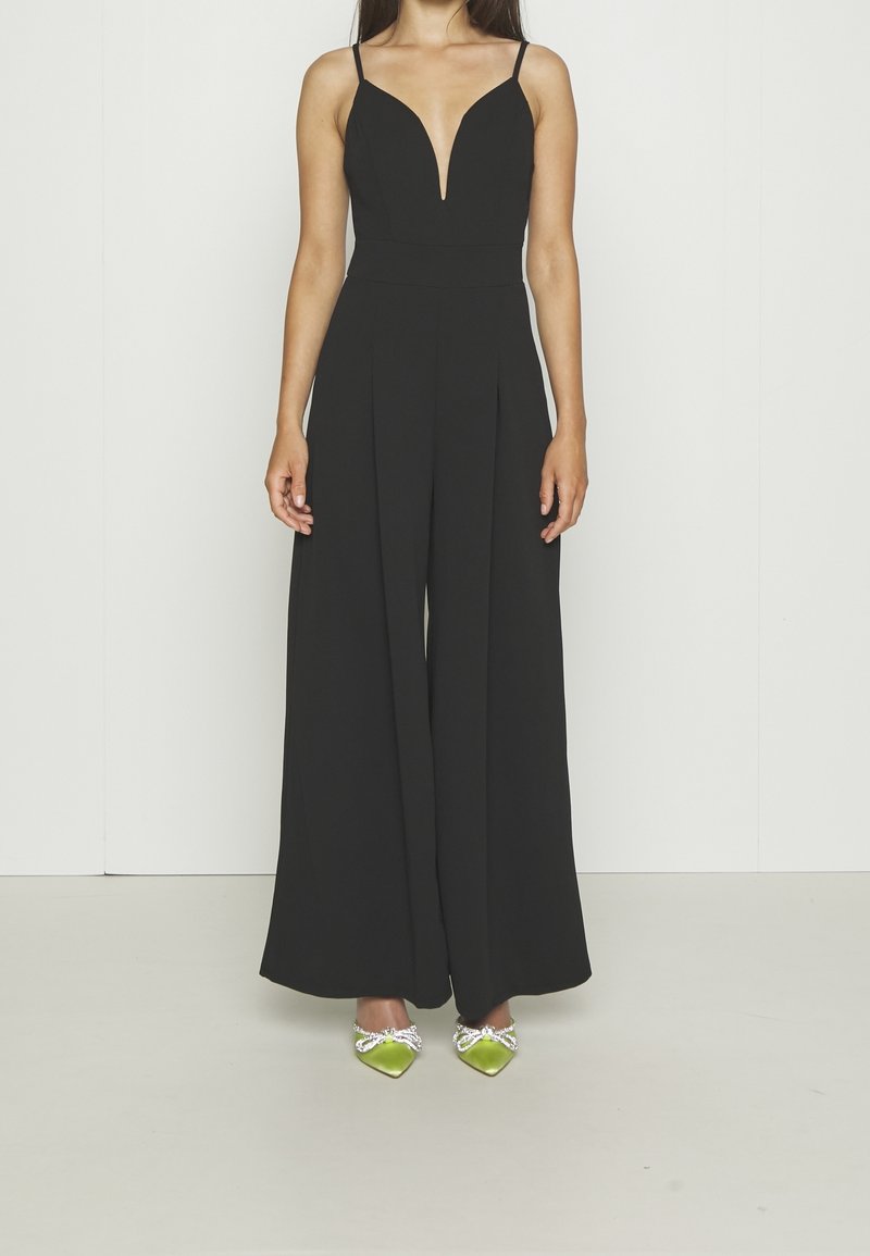 Femme portant une combinaison noire sans manches à jambes larges et col en V profond, associée à des chaussures vertes pointues ornées de nœuds en cristal.