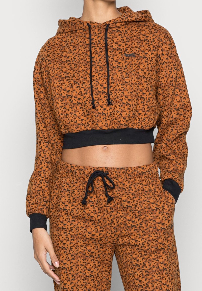 Femme portant un sweat à capuche court imprimé léopard orange et noir avec un pantalon assorti aux poignets et cordons noirs.