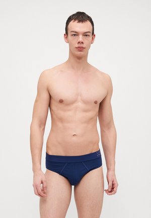 Jeune homme debout, regardant devant lui, portant un slip bleu marine sur un fond blanc uni, affichant une expression neutre.