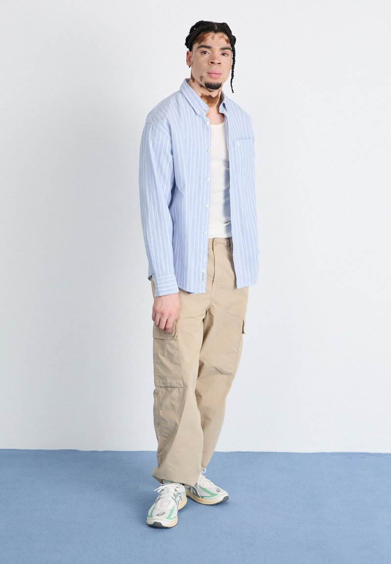 Camisa de botones a rayas en azul claro y blanco, camiseta sin mangas color crema, pantalones cargo beige y zapatillas blancas con detalles en verde. Diseño simple, ajuste casual.