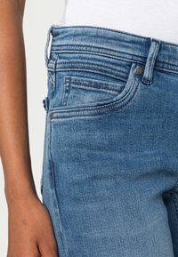 Jeans en denim bleu avec une coupe ajustée, comportant une disposition à cinq poches et des accents en rivets en cuivre. Couture visible et tissu texturé.