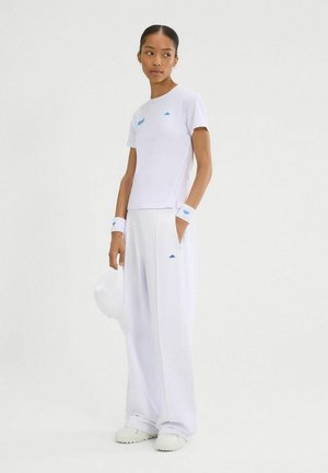 Camiseta blanca de manga corta con logo azul, combinada con pantalones blancos a juego y muñequeras. El modelo sostiene una gorra blanca.