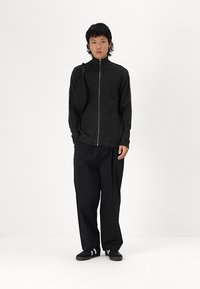 Schwarze Zip-Jacke aus strukturiertem Stoff, getragen über locker sitzenden schwarzen Hosen. Eine kleine schwarze Tasche ist über eine Schulter geschlungen.