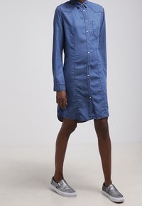 Abito camicia in denim con colletto, bottoni frontali, maniche lunghe, tasca sul petto e orlo curvo. Abbinato a scarpe slip-on argentate.
