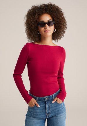 Haut rouge à manches longues côtelé avec un col bateau, associé à un jean bleu taille haute. Le haut présente des accents de boutons aux poignets.