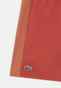 Calções em tecido bicolor vermelho e castanho com textura suave e pequeno logótipo de crocodilo verde bordado na bainha.