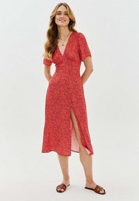 Červené květinové šaty s krátkými rukávy, hlubokým výstřihem do V a rozparkem vpředu. Lehká látka s volným střihem, se živým vzorem.