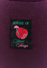 Fialová látka s černou etiketou. Na etiketě je nápis "DÁVEJ POZOR NA" zelenou barvou, červené jablko s bílými tečkami, růžová houbu a nápis "Sladké věci" růžovým kurzívou.