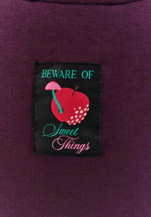 Fioletowa tkanina z czarną metką. Metka zawiera napis "UWAŻAJ NA" w zielonym, czerwone jabłko z białymi kropkami, różowy grzyb oraz "Słodkie Rzeczy" w różowym kursywie.