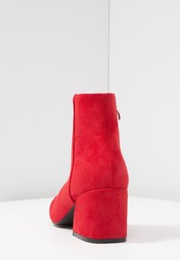 Czerwone zamszowe botki za kostkę z masywnym czarnym obcasem, charakteryzujące się gładką fakturą i eleganckim designem. Widok z tyłu ukazuje minimalistyczną sylwetkę.
