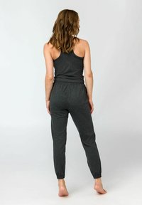 Frau steht barfuß mit dem Rücken zur Kamera, trägt ein ärmelloses, dunkelgraues Oberteil und passende Jogginghosen vor einem schlichten, hellen Hintergrund.