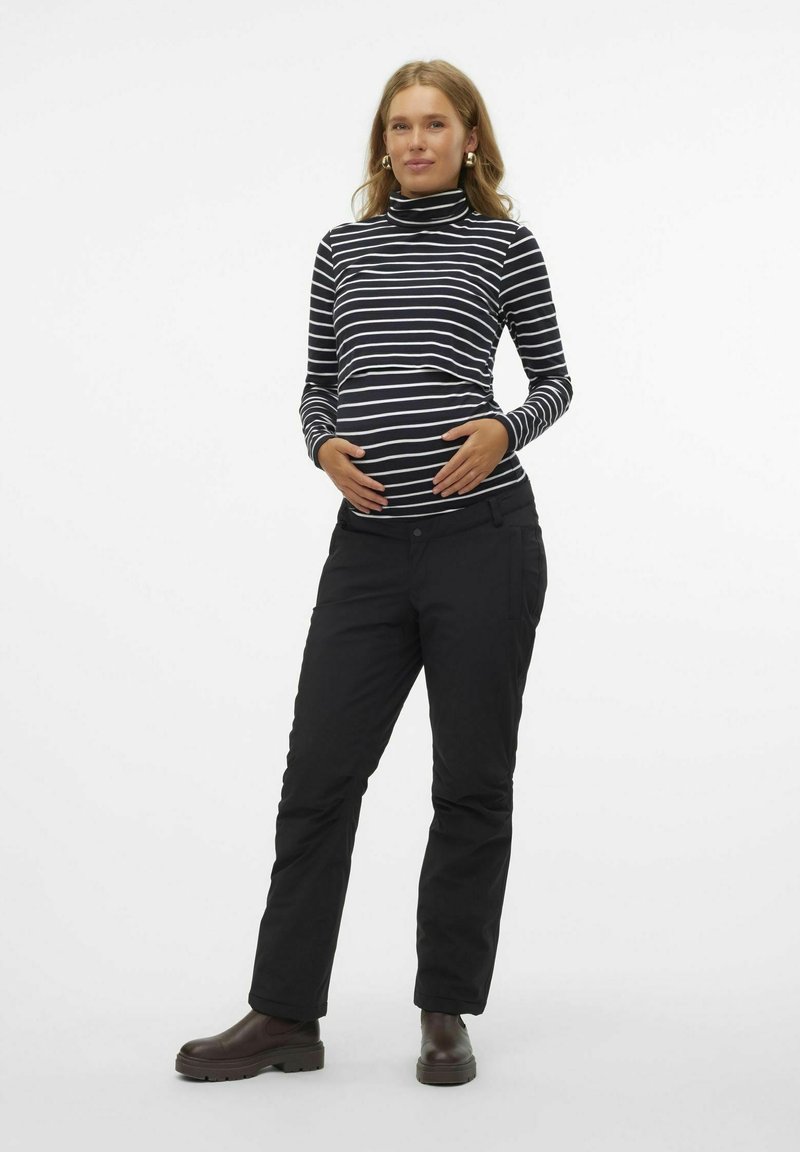 Pantalon noir et un haut à col roulé noir et blanc rayé, porté par une personne avec un ventre de femme enceinte visible, se tenant dans un cadre neutre.