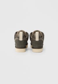 Σκούρα ελιάς πράσινα σουέντ αθλητικά παπούτσια με μπεζ κορδόνια και επένδυση από shearling, εμφανίζονται από πίσω σε ανοιχτό γκρι φόντο.