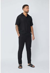 Polo noir à manches courtes avec un col plat, associé à un pantalon fuselé assorti et des sandales noires, mettant en valeur un tissu lisse et une coupe décontractée.