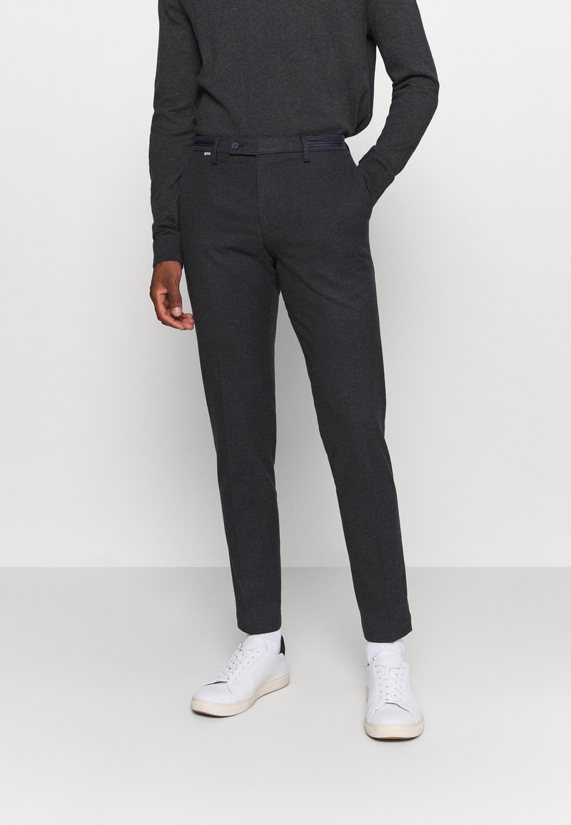 Personne portant une chemise à manches longues gris foncé, un pantalon de costume assorti et des baskets blanches, debout devant un fond uni clair.