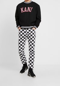 Felpa nera con scritta "KANI" in bianco e rosso, abbinata a pantaloni a scacchi in bianco e nero. Sneakers nere completano l'outfit.